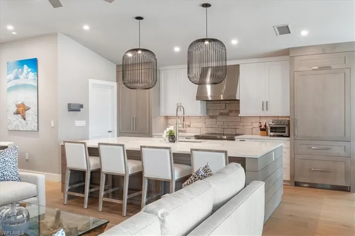 Kitchen featuring wall chimney exhaust hood, light wood-style floors, tasteful backsplash, a kitchen island with sink, and light stone counters