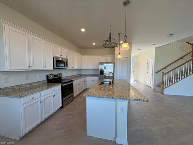 Kitchen featuring appliances with stainless steel finishes, white cabinetry, decorative light fixtures, light stone countertops, and a kitchen island with sink