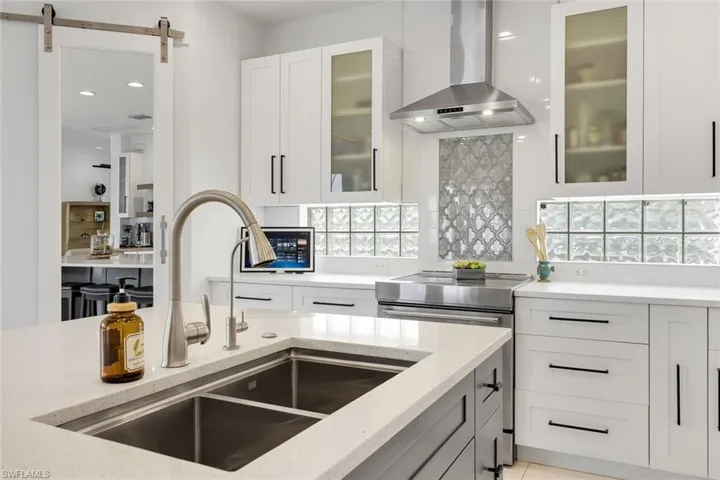 Stainless steel double basin sink with a pull-down faucet, white perimeter cabinetry with dark hardware, glass block window accents, and a stainless steel range with a coordinating vent hood