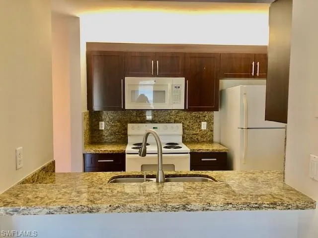 Kitchen with dark wood finish cabinetry, white appliances, light stone counters, a peninsula, and decorative backsplash