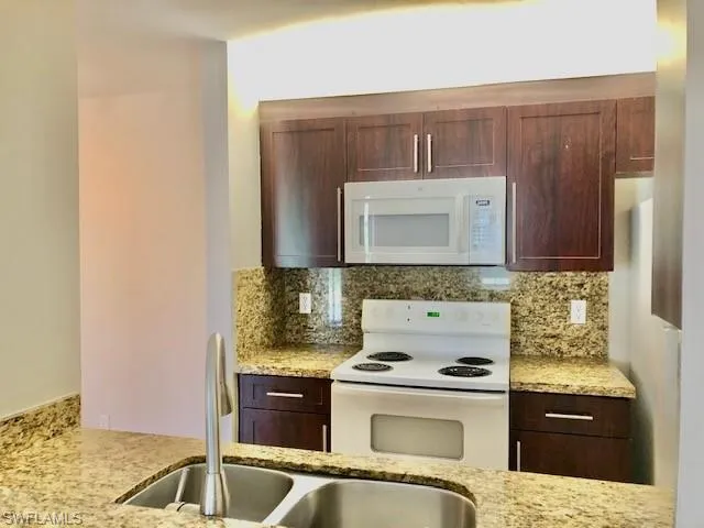 Kitchen with white appliances, light stone countertops, dark wood finish cabinets, and backsplash