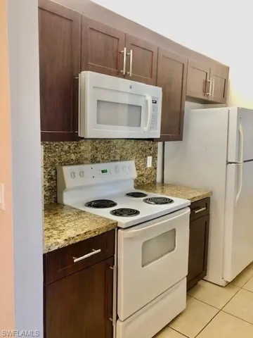 Kitchen with white appliances, dark wood finish cabinetry, backsplash, and light stone countertops