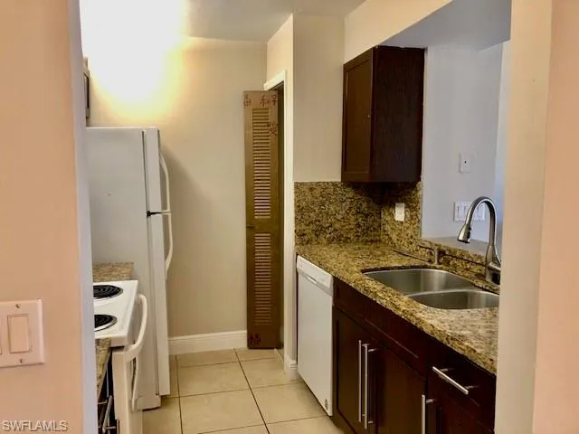 Kitchen with dark wood finish cabinetry, light stone counters, white appliances, light tile patterned floors, and tasteful backsplash