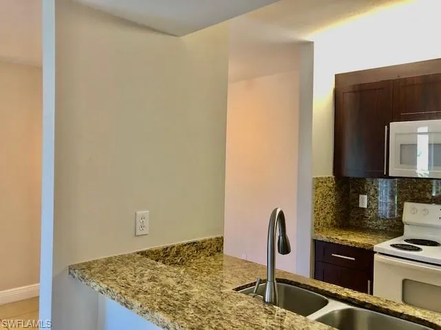 Kitchen with dark wood finish cabinets, white appliances, light stone countertops, tasteful backsplash, and a peninsula