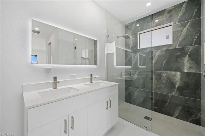Full bath with double vanity, a marble finish shower, a textured wall, and recessed lighting