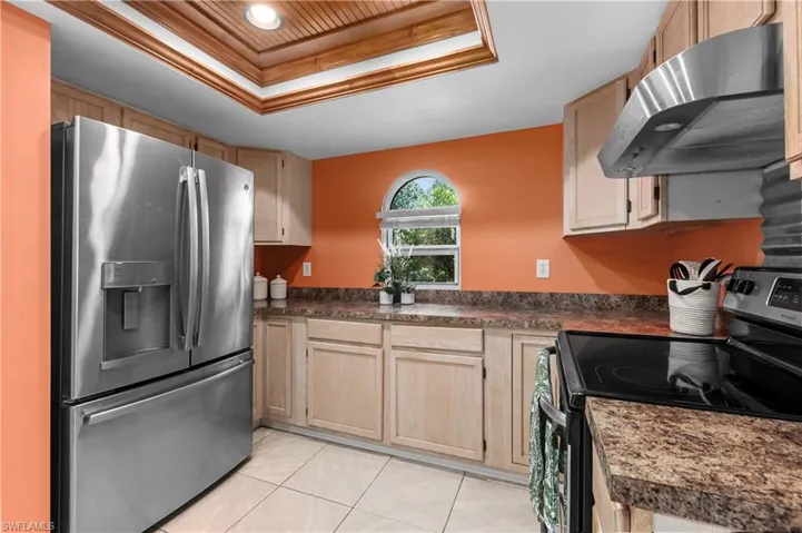 Kitchen featuring electric range oven, a tray ceiling, stainless steel fridge, under cabinet range hood, and light brown cabinetry