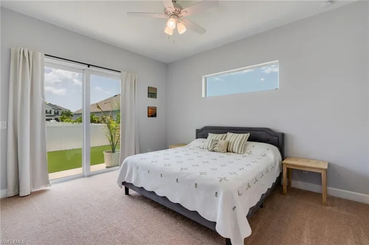 Carpeted room featuring a ceiling fan with integrated lighting, a horizontal transom window, and a sliding glass door leading to a backyard with artificial turf and a privacy fence