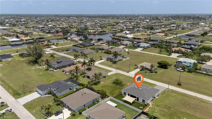 Aerial perspective highlighting a single-story home with a dark shingle roof, a paved driveway, and a fenced backyard featuring a shade sail structure