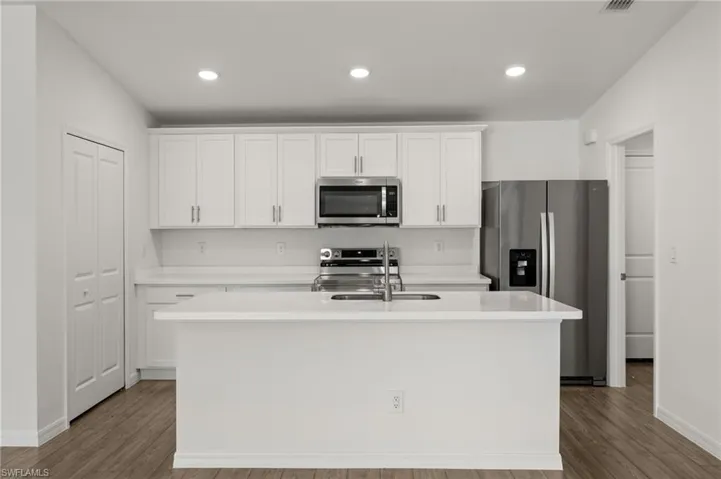 Kitchen with a kitchen island with sink, stainless steel appliances, wood finished floors, white cabinetry, and light countertops
