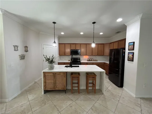 Kitchen with black appliances, ornamental molding, brown cabinets, light stone counters, and a kitchen island with sink