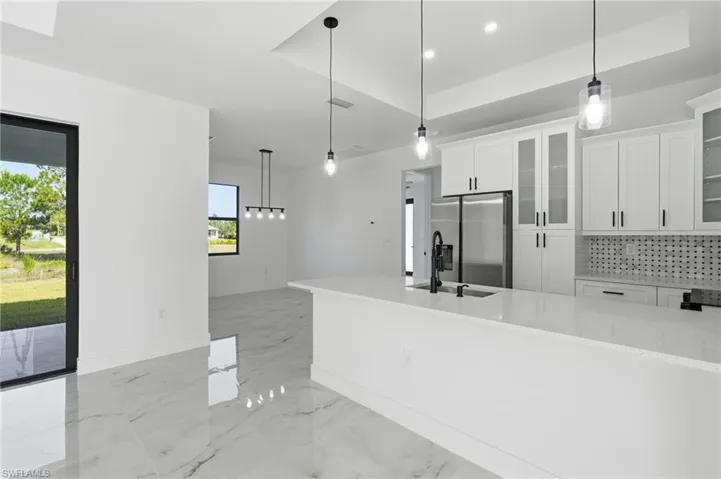 Kitchen island with a built-in sink and matte black faucet, white shaker cabinetry, stainless steel refrigerator, patterned tile backsplash, and large format tile flooring