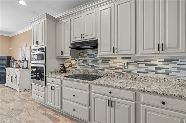 Kitchen featuring black electric stovetop, under cabinet range hood, tasteful backsplash, ornamental molding, and stainless steel double oven