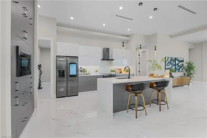 Kitchen featuring black microwave, stainless steel refrigerator with ice dispenser, a center island with sink, wall chimney range hood, and light tile patterned floors