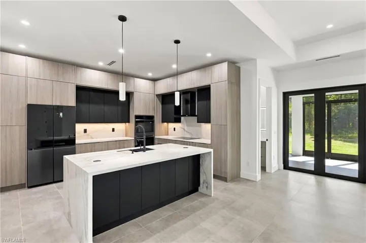 Kitchen featuring flat-panel cabinetry in contrasting wood-finish and matte black, stone-finish countertops, full-height stone-finish backsplash, stainless steel range hood, and a flush-mounted cooktop