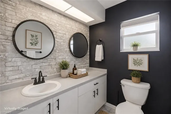 Light-toned brick accent wall, white vanity with integrated sink, dark-toned walls, double round mirrors, and a window with frosted glass
