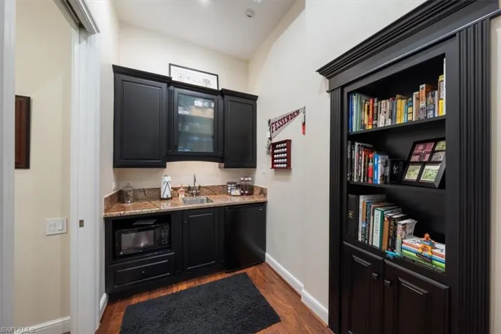Indoor wet bar featuring dark cabinetry, dark wood-style flooring, black appliances, and light stone counters