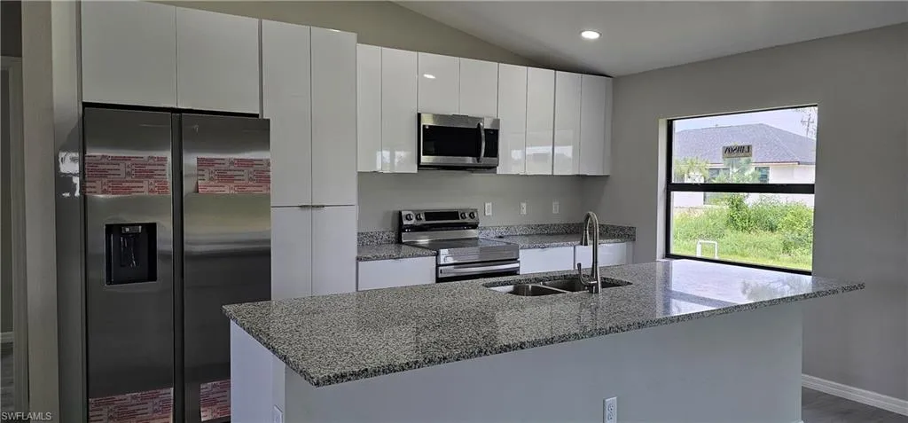 Kitchen with appliances with stainless steel finishes, white cabinetry, vaulted ceiling, a kitchen island with sink, and light stone countertops