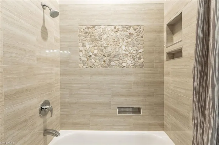 Same guest bathroom with tub, shower, stones on the wall, and storage places to store soap & other utensils.