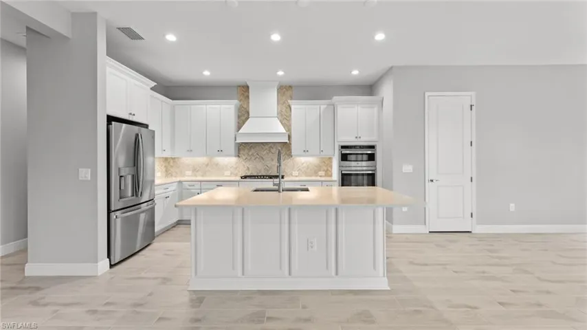 Kitchen featuring tasteful backsplash, custom exhaust hood, stainless steel appliances, a kitchen island with sink, and white cabinetry