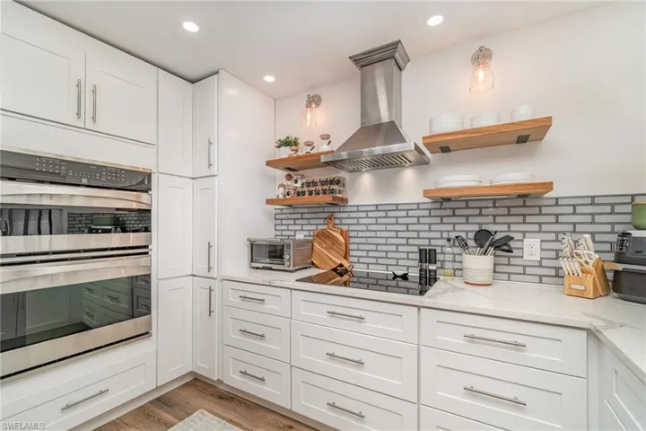 Kitchen featuring stainless steel double oven, wall chimney exhaust hood, open shelves, decorative backsplash, and recessed lighting