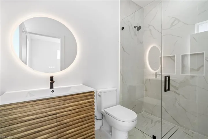 Contemporary bathroom featuring a floating wood-finish vanity with an integrated sink, a backlit circular mirror, and a glass-enclosed shower with marble-look tile surrounds and built-in niches