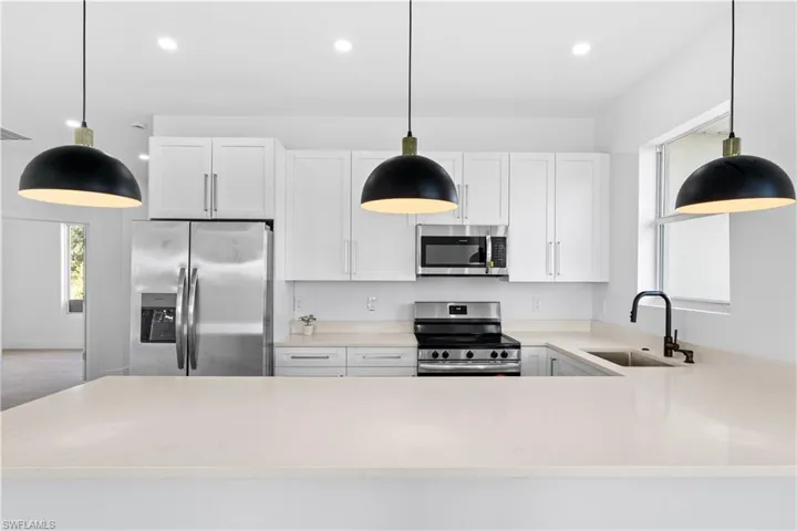 Modern kitchen featuring white shaker cabinetry, stainless steel appliances, light-toned countertops, a black wall-mounted faucet, and three contemporary pendant lights