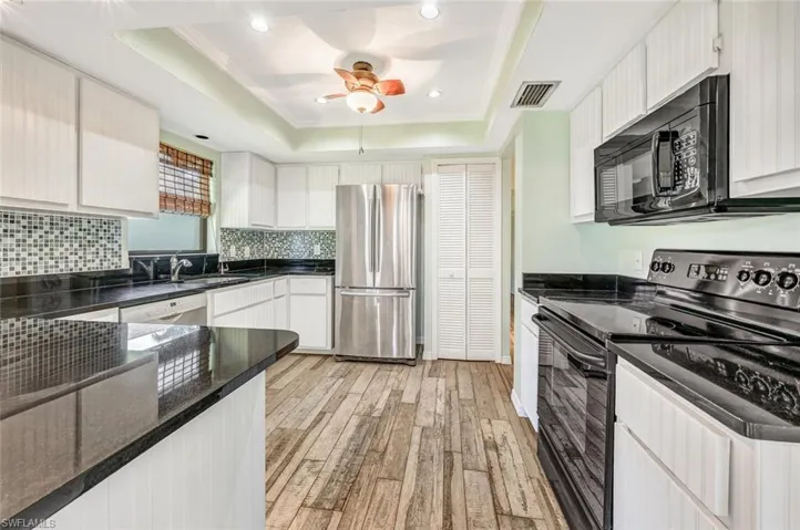 Kitchen featuring light hardwood / wood-style floors, dark stone countertops, a tray ceiling, black appliances, and ceiling fan