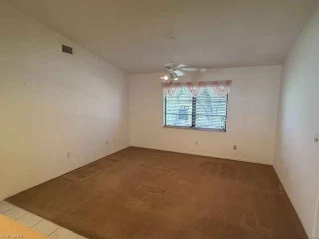 Spacious interior room featuring a large window with blinds, ceiling fan with integrated lighting, and neutral-toned wall-to-wall carpeting