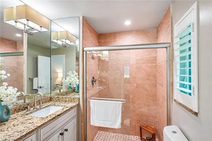 Shower with tile surround and glass enclosure, vanity with stone countertop, mounted mirror, and plantation shutters