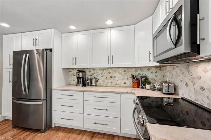 White cabinetry with brushed nickel hardware, stainless steel appliances, patterned tile backsplash, light-toned countertops, and wood-finish flooring