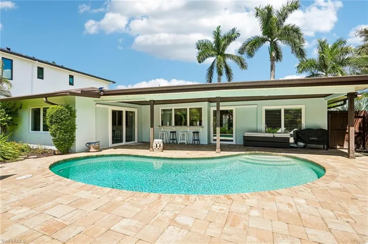 Resort-style backyard featuring a freeform swimming pool, extensive paver decking, a covered patio, and mature palm trees