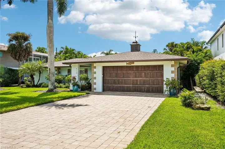 Exterior featuring a paver driveway, wood-finish garage door, cupola with weathervane, and mature tropical landscaping