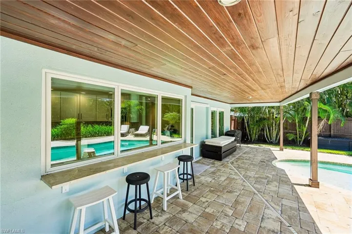 Covered patio featuring a wood-panel ceiling, stone paver flooring, and a built-in outdoor bar with a pass-through window to the interior