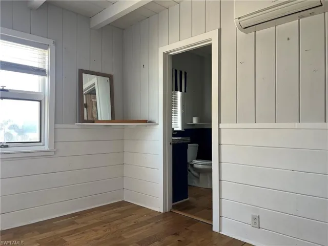 Current  view primary bedroom with wood walls, beamed ceiling, a wall mounted AC, and dark hardwood / wood-style flooring with attached bathroom