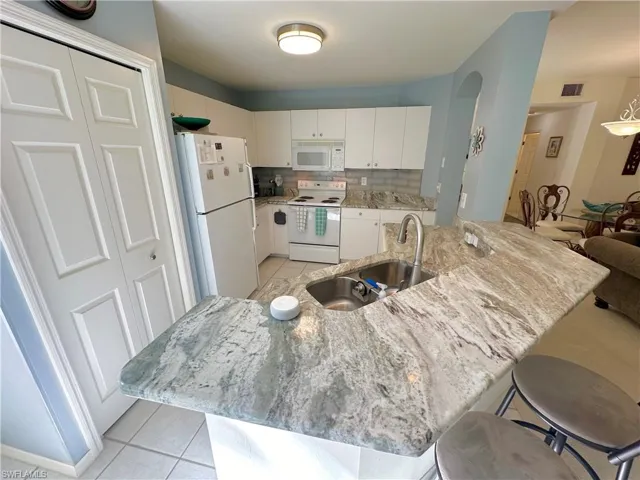 Kitchen with light tile patterned floors, white appliances, a kitchen breakfast bar, white cabinetry, and light stone counters