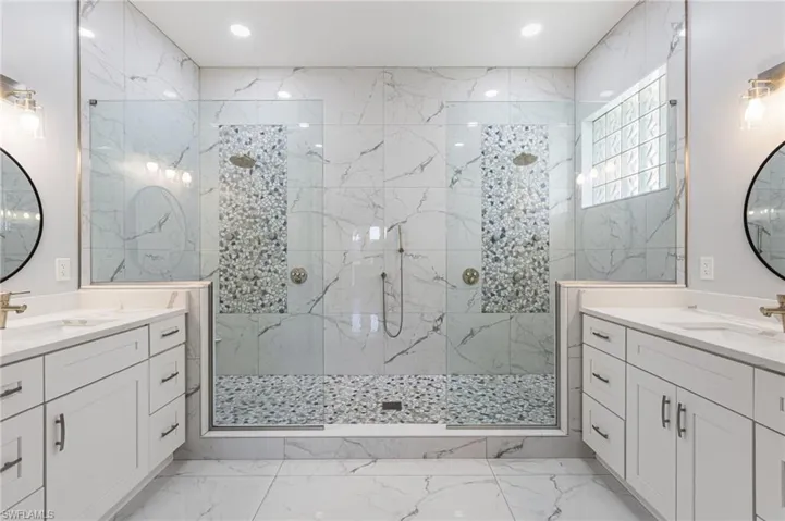 The completely reimagined primary bathroom feels more like a private spa retreat, featuring his-and-hers quartz-topped vanities, a luxurious shower with dual heads, body sprays, and a handheld wand, all enclosed in seamless glass. Floor-to-ceiling porcelain walls and a striking listello add a designer touch, while custom cabinetry with pull-out drawers delivers impressive storage with style to spare.