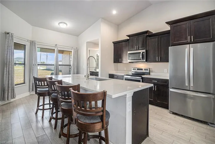 Kitchen with appliances with stainless steel finishes, vaulted ceiling, dark brown cabinetry, light stone counters, and a center island with sink