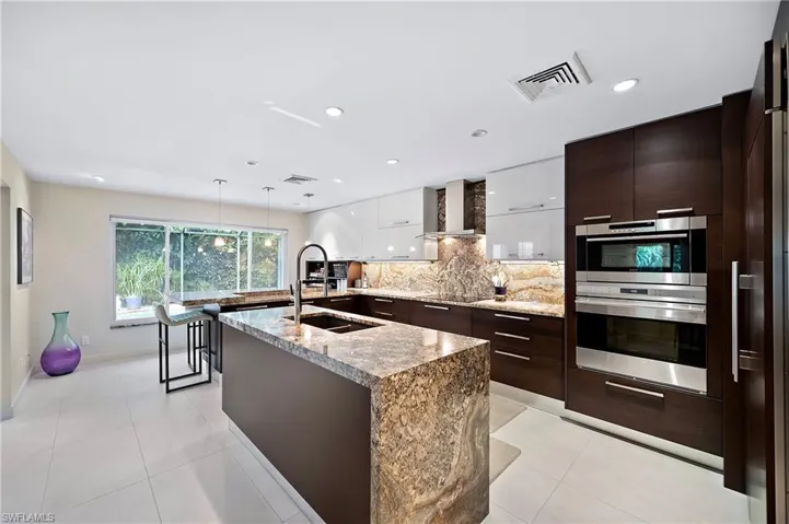 Kitchen featuring tasteful backsplash, wall chimney range hood, a center island with sink, double oven, and light tile patterned floors