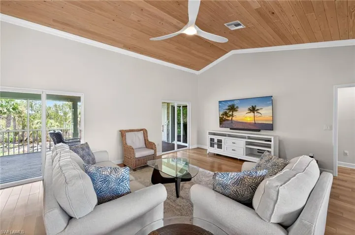 Spacious living area featuring a vaulted wood-paneled ceiling, light wood-finish flooring, white crown molding, and direct access to an outdoor deck through sliding glass doors
