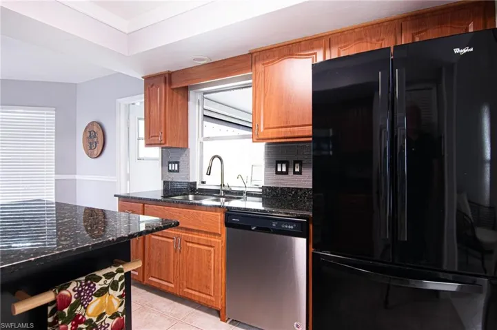 Kitchen with freestanding refrigerator, dark stone countertops, stainless steel dishwasher, and brown cabinetry