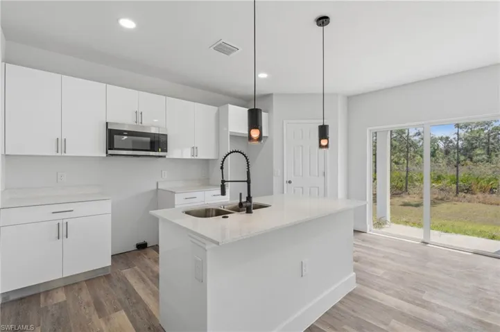 Kitchen featuring white cabinets, pendant lighting, stainless steel microwave, light wood-style floors, and a kitchen island with sink