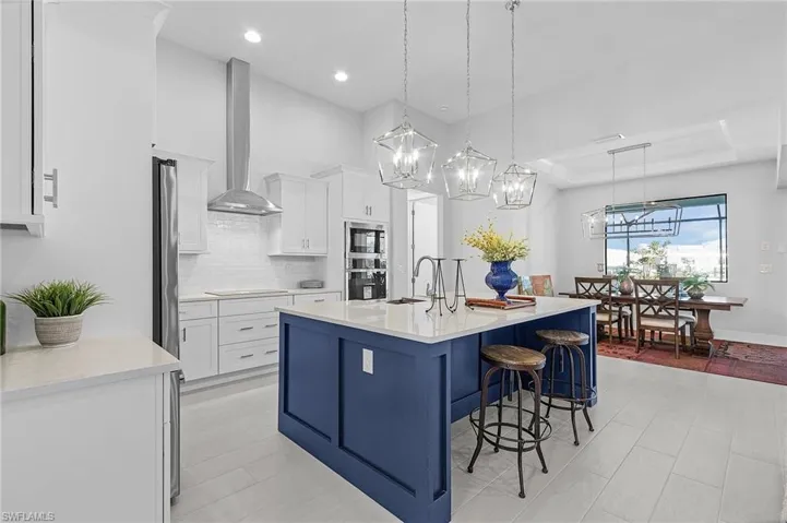 Kitchen featuring wall chimney range hood, hanging light fixtures, a tray ceiling, a center island with sink, and white cabinets
