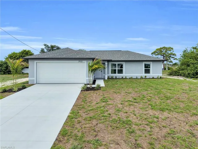 Newly constructed residence featuring a light gray exterior with white trim, an attached garage, a paved driveway, and a front landscaping package with mature palms
