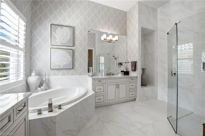 Bathroom featuring stone-finish flooring and wall tiling, a soaking tub with stone-finish surround, a glass-enclosed shower, built-in vanity cabinetry, and plantation shutters