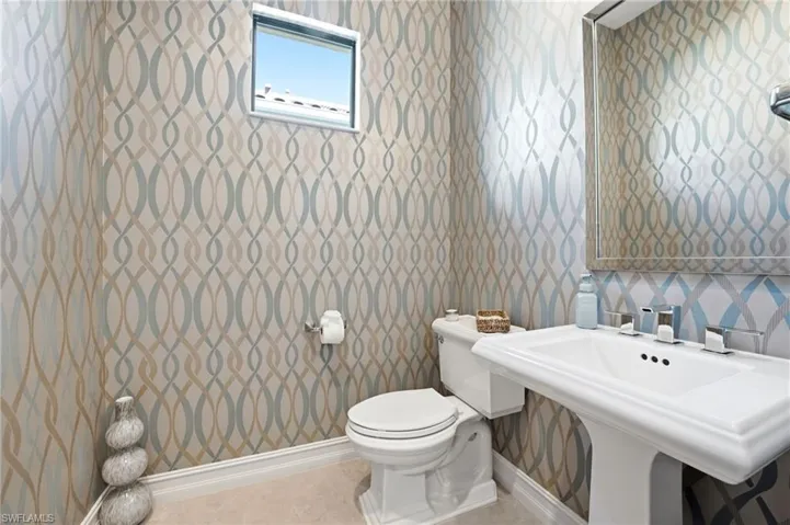 Powder room featuring patterned wallpaper, a pedestal sink with a polished chrome faucet, a rectangular framed mirror, and a window