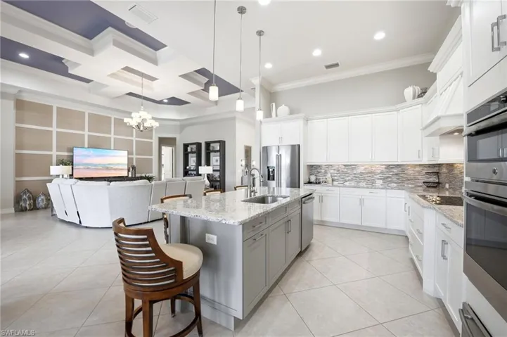 Kitchen featuring a central island with stone countertop and stainless steel sink, custom white shaker-style cabinetry, a multi-textured stone tile backsplash, and built-in stainless steel double ovens