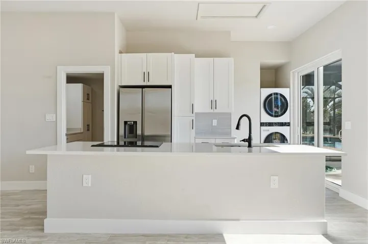 Kitchen with a kitchen island with sink, stainless steel refrigerator with ice dispenser, white cabinets, light stone countertops, and stacked washer and dryer - Virtually Edited Image
