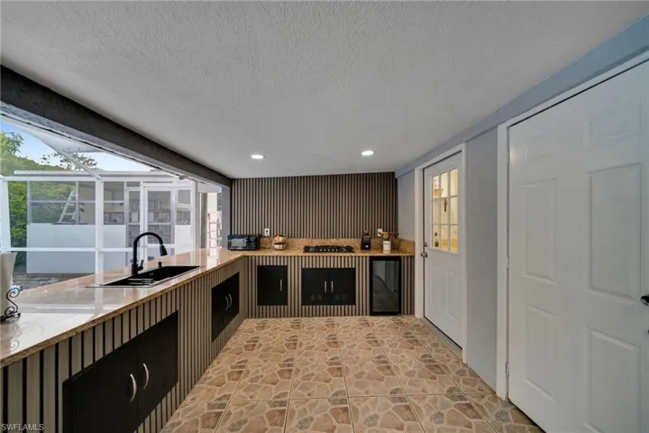 Outdoor kitchen featuring a stone-look tiled floor, an integrated sink with a matte black faucet, light-toned countertops, built-in cabinetry, and a screened enclosure