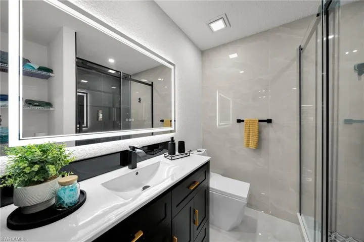 Contemporary bathroom featuring a black vanity with gold-tone hardware, a white countertop with an integrated sink, an illuminated mirror, a glass-enclosed shower, and large format wall tiles