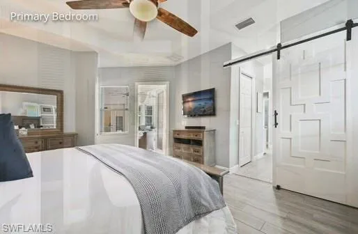 Bedroom with a barn door, ceiling fan, ensuite bath, and light hardwood / wood-style flooring